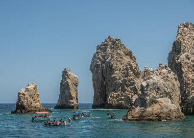Meksika, Cabo San Lucas - 16 Temmuz 2023: gri-kahverengi rshort ve okyanustan yükselen uzun oktavlı kayalar, mavi gökyüzünün altındaki yarımadanın sonu. Birkaç küçük turist gezi teknesi