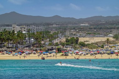 Meksika, Cabo San Lucas - 16 Temmuz 2023. Constituyentes insanları, şemsiyeleri ve Tabasco barını kumsala götürüyor. Mavi gökyüzü ve dağların altındaki Cascadas de Baja Kulüp binası.