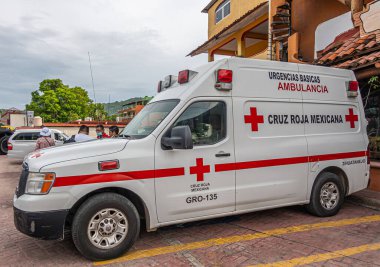 Zihuatanejo, Meksika - 18 Temmuz 2023: Rıhtımın sonunda gri bulutların altında park edilmiş kırmızı-beyaz ambulans. Arkaplan olarak sarı bina cephesi. İnsanlar ve yeşil yapraklar