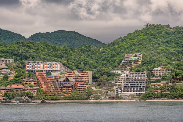 Zihuatanejo, Meksika - 18 Temmuz 2023: Kumlu doğu kıyı şeridi. Tatil köyleri, gri bulutların altındaki yeşil ormanlık tepeyi biçimlendirmek için teraslarda renkli mimari yapılar oluşturur.. 