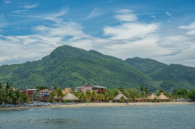 Zihuatanejo, Meksika - 18 Temmuz 2023: Playa La Madera, mavi bulutlar altında yeşil ormanlık tepelerin önünde gölgelik yapmak için saman kaplı dairesel açık duvar kulübeleriyle. Tekneler ve evler