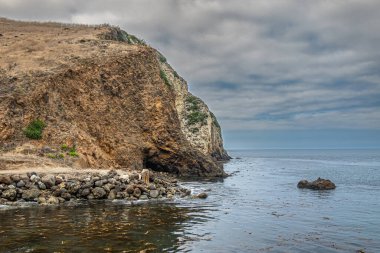 Santa Cruz Adası, CA, ABD - 14 Eylül 2023: Kahverengi-beyaz kayalık uçurum Akrep Anchorage 'ın hemen kuzeyinde ve sınırında mavi bulutlar altında. Mavi okyanus suyu, bazı yeşil alanlar.