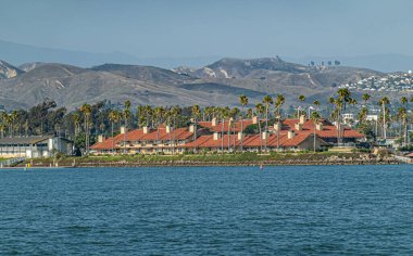 Ventura, CA, ABD - 14 Eylül 2023: Ventura Harbor Village Alışveriş Merkezi binası mavi okyanus suyunun arkasında kırmızı çatılı. Yeşil palmiye ağaçları, mavi gökyüzü ve arkadaki dağ sıraları