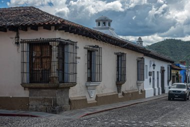 Guatemala, La Antigua - 20 Temmuz 2023: Metal ızgaralar Avenida Norte ve Calle Poniente 'nin köşesindeki pencereleri koruyor. Ağır mavi gri bulut manzarası. Sokakta arabalar. Arkadaki yeşil dağ.