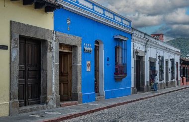 Guatemala, La Antigua - 20 Temmuz 2023 Calle Poniente. Giriş ve ızgara korumalı pencereli Gascon mavi cephesi. Sokak manzarası ve ağır mavi gri bulutlar. Arkadaki yeşil dağ.