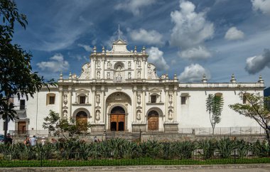 Guatemala, La Antigua - 20 Temmuz 2023: Beyaz taş San Jose Katedrali Plaza Belediye Başkanı, merkez meydanında, mavi bulutların altında. Önde kırmızı çiçekler ve yeşil yapraklar var.