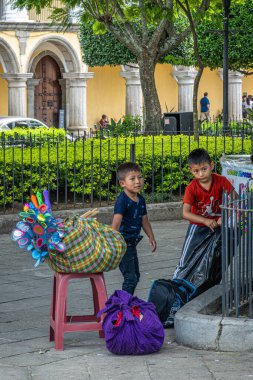 Guatemala, La Antigua - 20 Temmuz 2023: 2 küçük çocuk Plaza Mayor 'da aile mallarını satılığa çıkarıyor. Yeşil yaprakların arasında, Ulusal Sanat Müzesi 'nin sarı cephesi
