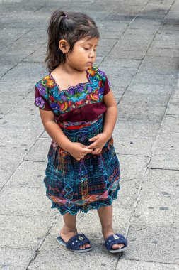 Guatemala, La Antigua - 20 Temmuz 2023: Kapanış, renkli geleneksel giysiler giymiş küçük bir kız üzgün bir yüzle Plaza Belediye Başkanı 'nın gri kaldırımına bakıyor.