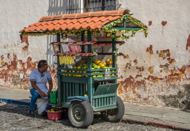 Guatemala, La Antigua - 20 Temmuz 2023: Erkek sokak meyve satıcısı yeşil el arabasıyla caddenin kenarında oturuyor. Arkaplan olarak paslı lekeli bej duvar
