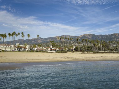 Santa Barbara, CA, ABD - 27 Ocak 2024: Stearns Wharf bakış açısı. Doğu Cabrillo Bulvarı 'nın ilk üç bloğu kumlu doğu sahilinin arkasında, mavi bulutların altında. Beyaz cepheler, yeşil palmiye ağaçları, dağlar,