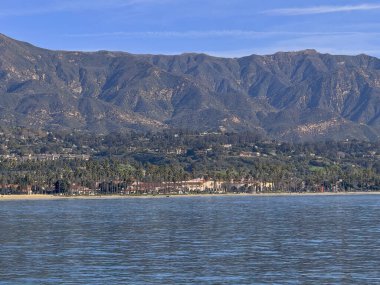 Santa Barbara, CA, ABD - 27 Ocak 2024: Hilton Beachfront Resort, East Beach 'in arkasındaki yeşil palmiye ağaçlarında geçiyor. Mavi bulutların altında bir arkaplan gibi dağ aralığı