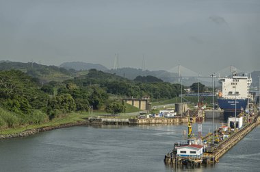 Panama Kanalı, Panama - 24 Temmuz 2023: Miraflores 'in sol odaları açık mavi gökyüzü altında kuzeye gitmek için kilitlenir. CMA CGM konteyner gemisi. Ufukta orman dağları olan yüzüncü köprü