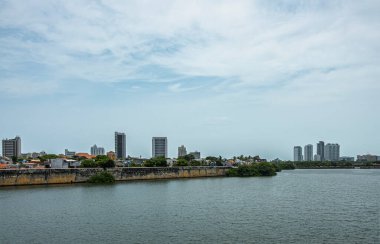Cartagena, Colombia - July 25, 2023: Looking over bay water at old town from Puente Roman bridge. Historic reinforced shore ramparts under light blue cloudscape. Few tall buildings clipart