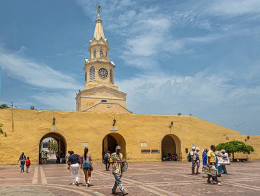 Cartagena, Colombia - July 25, 2023: Torre del Reloj and gate in remaining part of ramparts into old town center seen from Plaza de los Coches under blue cloudscape. Pedestrians and street vendors clipart