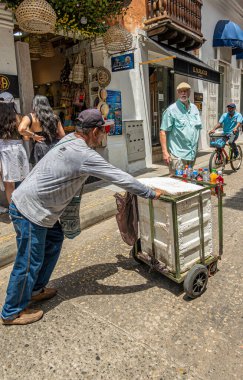 Cartagena, Kolombiya - 25 Temmuz 2023: Küçük el arabasında köpük kutusu olan sokak satıcısı Carrera 3 boyunca şişe soda ve su satıyor. Diğer yayalar ve küçük işletmeler