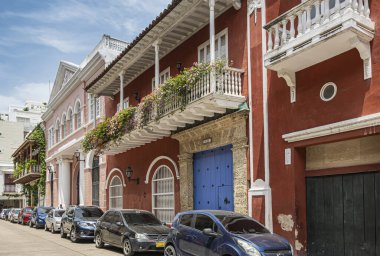 Cartagena, Colombia - July 25, 2023: Colonial architecture in Calle de la Inquisicion with parked cars under blue cloudcape. Balconies with flowers and green foliage clipart