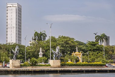 Cartagena, Kolombiya - 25 Temmuz 2023: Arkasında yeşil Centenario Parkı olan eski kasaba rıhtımının sonundaki Double Pegasus heykelleri. Mavi bulutların altında sarı anıtsal bir kapı var. Yüksek beyaz bina