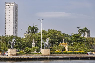 Cartagena, Colombia - July 25, 2023: Double Pegasus statues at end of old town dock with green Centenario Park behind. Yellow monumental gate into the it under blue cloudscape. Tall white building clipart