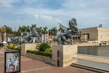 Cartagena, Kolombiya - 25 Temmuz 2023: Eski kasaba rıhtımının sonundaki Double Pegasus heykelleri ve mavi bulutların altındaki kongre merkezi. Yoyo Jeans için palmiye ağaçları ve poster.