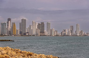 Cartagena, Kolombiya - 25 Temmuz 2023: Calle 33 'ün Avenida Santander ile buluştuğu yerden, Bocagrande yarımadasının NW tarafında yüksek binalar silueti. Önde kayalık kıyı, mavi su, bulutlu manzara