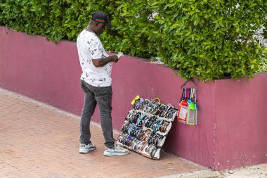 Cartagena, Colombia - July 25, 2023: Central Bocagrande Carrera 2. Street vendor sells sunglasses. Green foliage above pink wall at his back clipart
