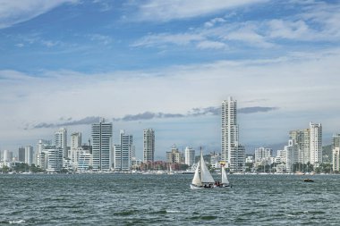 Cartagena, Kolombiya - 25 Temmuz 2023: Mavi bulutların altındaki beyaz uzun binaların önündeki yat limanı. Ön tarafta bir yat var.