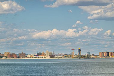 New York, New York, ABD - 1 Ağustos 2023: Coney Island sahilinde kırmızı kulesi olan Maimonides Parkı mavi bulutların altında. Önde gri okyanus suyu var. Kahverengi ve sarı binalar arkasında