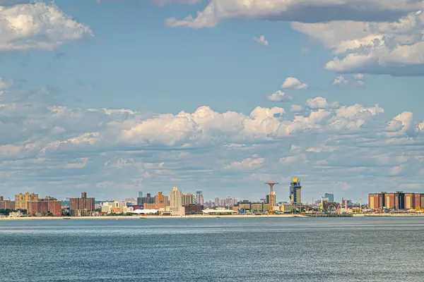 stock image New York, NY, USA - August 1, 2023: Maimonides Park with red tower on Coney Island beach under blue cloudscape. Gray ocean water up front. Brown and yellow buildings behind