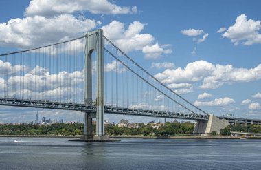 New York, New York, ABD - 1 Ağustos 2023: Verrazzano-Narrows Köprüsü E tarafı Fort Hamilton 'a iniyor. Ufukta mavi bulutların altında Manhattan yüksek binaları olan yeşil kıyı şeridi