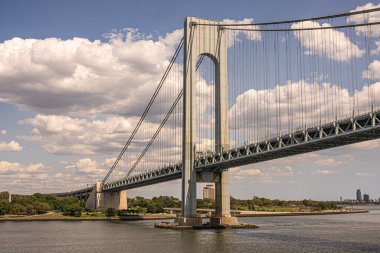 New York, NY, USA - August 1, 2023: N view, Verrazzano-Narrows Bridge E side half and landing at Fort Hamilton. Green coastline with tall buildings on horizon under blue sky clipart