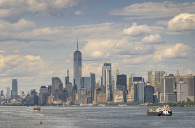 New York, New York, ABD - 1 Ağustos 2023: Bir Dünya Ticaret Merkezi ve Manhattan 'ın güneyindeki batarya parkının arkasındaki diğer finans bölgesi gökdelenleri mavi bulutlar altında. Nehirdeki tekneler