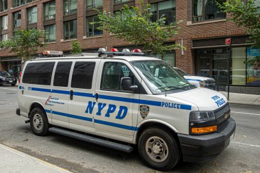 New York, New York, ABD - 3 Ağustos 2023: New York Polisi mavi-beyaz Chevrolet minibüsü yolda. Yeşil yapraklar ve tuğla ön cephe arkada.