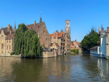 Brugge, Flanders, Belçika - 25 Haziran 2024: Rozenhoedkaai 'de görülen mavi gökyüzünün altında tarihi binalar ve Dijver kanalı üzerinde Halletoren,