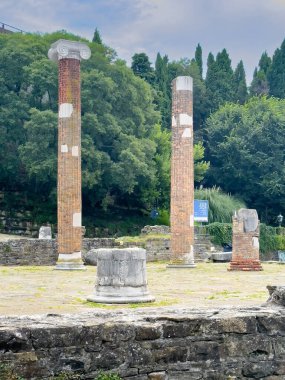 Trieste, Italy - June 27, 2024: Foro Romano, Roman Forum Archeological Park, a ruin of pillars short and tall, in front of Saint Justus Castle under a blue cloudscape. clipart