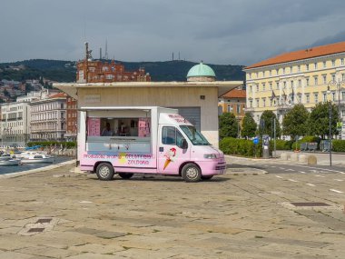 Trieste, İtalya - 27 Haziran 2024: Pembe ve beyaz renkli Gelateria Zoldana dondurma kamyonu sahil yolunda, arkada deniz suyu üzerinde küçük motorlu tekneler