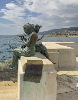 Trieste, İtalya - 27 Haziran 2024: Le Ragazze di Trieste heykeli Scale Reale 'de iki kız çocuğu, Adriyatik Denizi' ne girdi. Ufukta evleri olan ormanlık bir tepe. 2. Dünya Savaşı 'ndan sonra Tirestine kızlarının göçünü kutluyoruz.