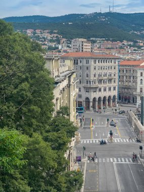 Trieste, İtalya - 27 Haziran 2024: Goldoni Meydanı 'ndaki Via Del Monte' den saray binaları, sokak seviyesindeki işleri ve üst kattaki ikametgahlarına bakmak. Ufukta orman tepeleri var. Araba ve yaya trafiği