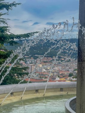 Trieste, Italy - June 27, 2024: Montuzza Fountain on top of Silvano Buffa Staircase, set in green park under blue cloudscape. Water sprays in the air while looking north over cityscape clipart