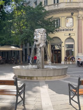 Trieste, Italy - June 29, 2024: Fountain of two-faced Janus, Fontana del Giano Bifronte, at entrance to via venti Settembre. Business facades and green foliage in back. benches in front clipart