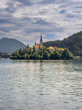 Bled Gölü, Slovenya - 28 Haziran 2024: Kutsal Bakire Meryem 'in Varsayımı Kilisesi kırmızı çatıları ve gri bulutlar altında kulesi olan küçük adada. Suda yıkanır.