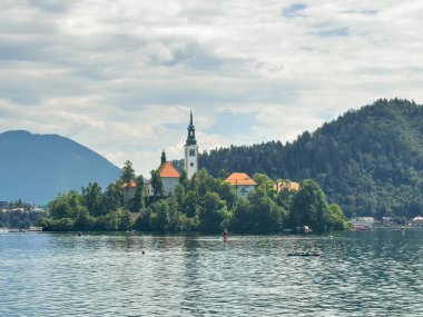 Bled Gölü, Slovenya - 28 Haziran 2024: Kutsal Bakire Meryem 'in Varsayımı Kilisesi kırmızı çatıları ve gri bulutlar altında kulesi olan küçük adada. Suda yıkanır. Ormanlık tepeler