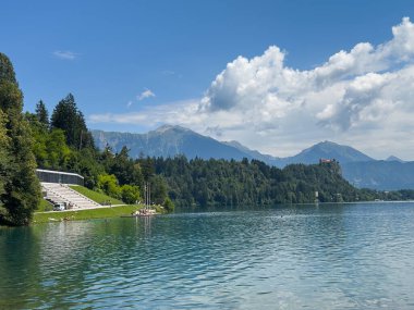 Bled Gölü, Slovenya - 28 Haziran 2024: Mavi bulutların altındaki ormanın arkasındaki kayanın arkasındaki şatoda kürek çekmek için bitiş çizgisi