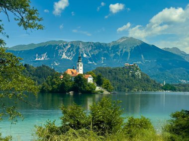 Bled Gölü, Slovenya - 28 Haziran 2024: Kutsanmış Bakire Meryem 'in Hükmü Kilisesi kırmızı çatıları ve kulesi olan küçük adada ve mavi bulutların altındaki kayanın arkasındaki kalede.