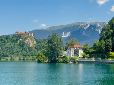 Bled Gölü, Slovenya - 28 Haziran 2024: Adora Oteli kıyıda, etrafı ağaç yapraklarıyla çevrili. Kale diğer tarafta, mavi gökyüzünün altında yeşil dağlar var..