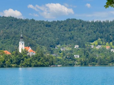 Bled Gölü, Slovenya - 28 Haziran 2024: Kutsanmış Bakire Meryem 'in Varsayımı Kilisesi' nin doğu tarafı kırmızı çatıları ve mavi bulutlar altında kulesi olan küçük adada. Kürekler suya. Ormanlık tepeler
