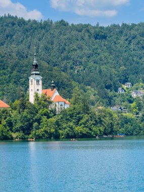 Bled Gölü, Slovenya - 28 Haziran 2024: Kutsanmış Bakire Meryem 'in Varsayımı Kilisesi' nin doğu tarafı kırmızı çatıları ve mavi bulutlar altında kulesi olan küçük adada. Kürekler suya. Ormanlık tepeler