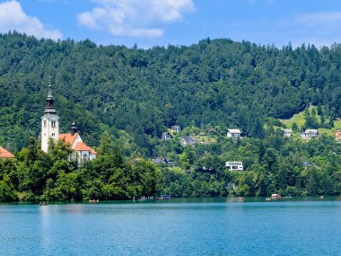 Bled Gölü, Slovenya - 28 Haziran 2024: Kutsanmış Bakire Meryem 'in Varsayımı Kilisesi' nin doğu tarafı kırmızı çatıları ve mavi bulutlar altında kulesi olan küçük adada. Kürekler suya. Ormanlık tepeler