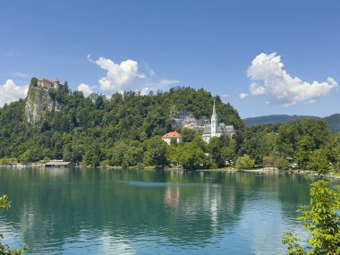 Bled Gölü, Slovenya - 28 Haziran 2024: St. Martin Kilisesi 'nin gri-beyaz mozaik çatısı mavi bulutların altında yeşil yapraklarla çevrili. Beyaz kayalıklar arkada, kalenin üstünde.