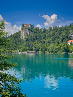 Slovenya 'nın Bled Gölü - 28 Haziran 2024: Kırmızı çatıları ile Şato ve müze, kayaların tepesinde, yeşil orman yaprakları ile çevrili mavi bulutlar altında. Doğu kıyısından görüldü