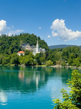 Bled Gölü, Slovenya - 28 Haziran 2024: St. Martin Kilisesi 'nin gri-beyaz mozaik çatısı mavi bulutların altında yeşil yapraklarla çevrili. Beyaz kayalıklar arkada. Geniş beyaz bölge ikametgahı ve yan taraftaki ofis.